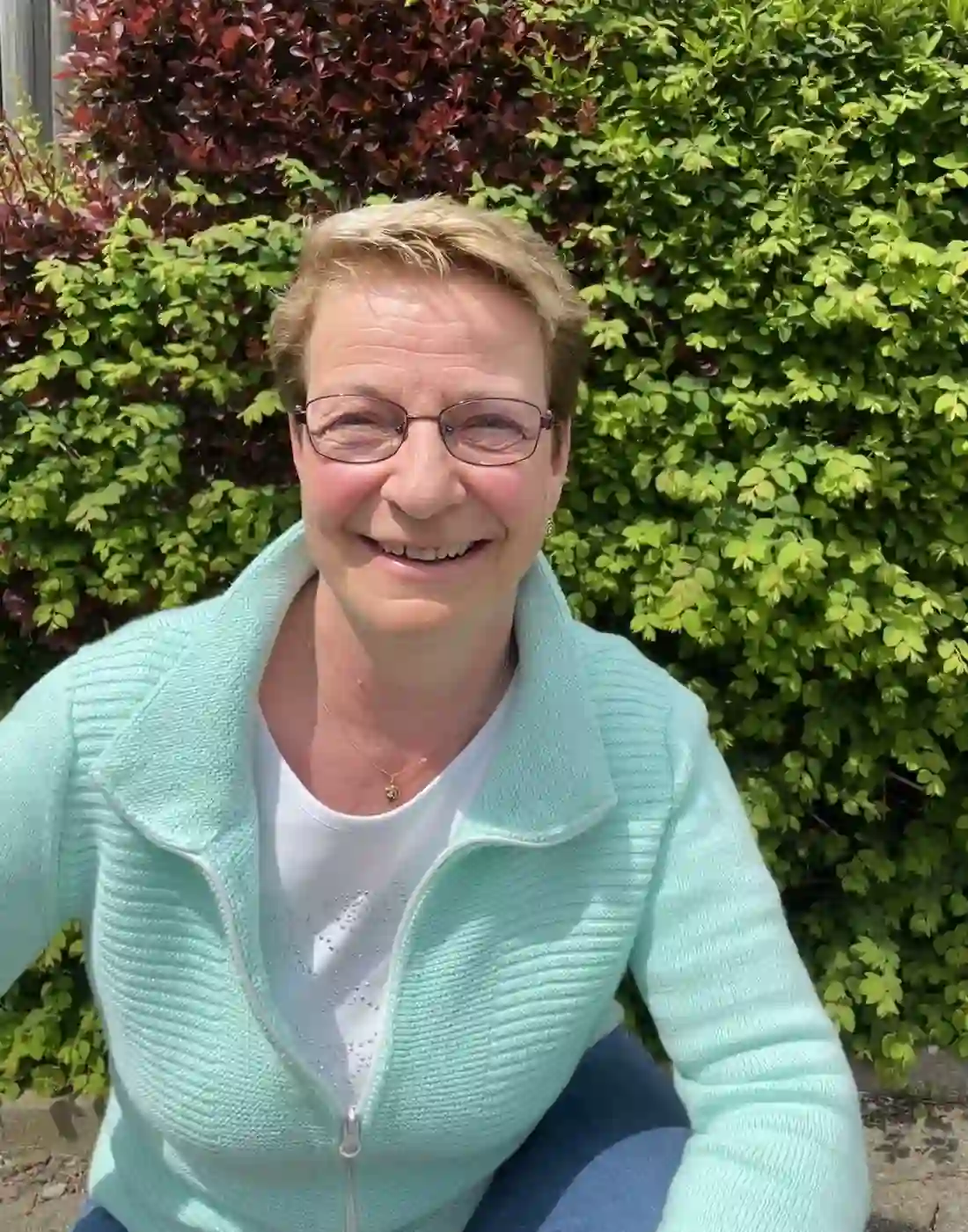 Porträt von Ulli Trüby in türkisfarbener Strickjacke vor einer grünen Hecke